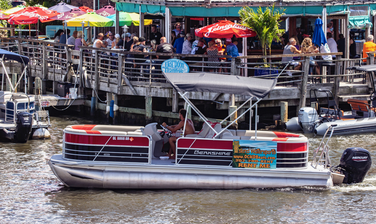 Group of people riding a black pontoon boat, with an ad for boat rentals on the side, near a dock.