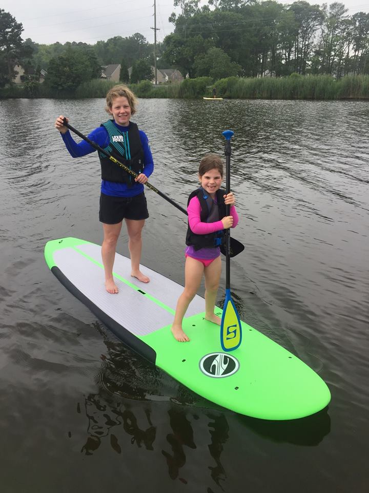 Two children riding a lime green paddle board while holding oars in the middle of the water.