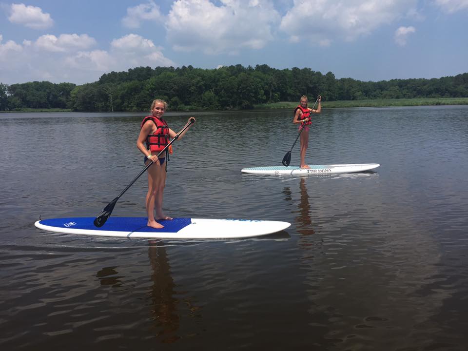 Two girls on two separate paddle boards in the middle of a lake with trees on the shore.